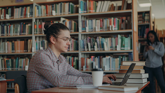 帅气的嬉皮士男学生坐在带现代笔记本电脑的轻便宽敞图书馆的桌子旁图片