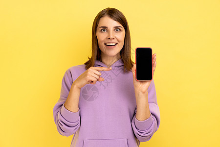 女人指着智能手机 空着展示宣传文字 微笑着看着相机 笑声享受帽衫紫色乐趣女性快乐黄色细胞女孩背景图片