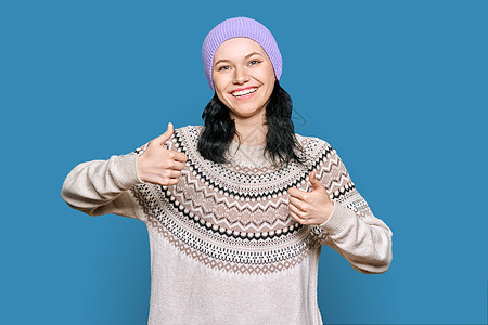 穿着冬季毛衣帽的快乐年轻女子 在蓝背景下举起拇指图片
