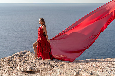 一个披着红色长裙 头发散乱的女孩站在海面上的岩石上 在背景中 大海和岩石 旅行的概念魅力幸福海滩海洋天后欲望手镯衣冠奢华女士图片