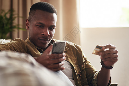 在网上看到喜欢的东西就下单吧 一个英俊的年轻人坐在家里的沙发上 使用智能手机和信用卡在线购物图片
