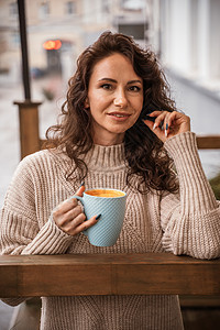一个中年妇女穿着米色毛衣 手里拿着一个蓝杯子 在阳台的街头咖啡厅里羊毛茶碗成人餐厅女孩温度衣服饮料冷冻女士图片