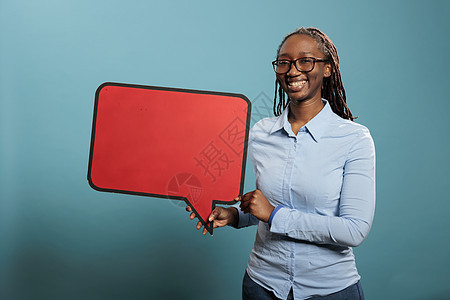 带着蓝底红纸板演讲泡泡的非洲自信和笑容女性在蓝色背景中 笑声成人木板空白横幅快乐广告牌说话广告讲话女士图片