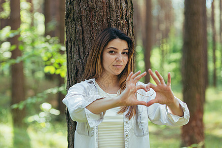 年轻美女志愿者在树旁的树林里做志愿者 微笑着看着镜头 用手和手指展示一颗心 这是热爱大自然的标志太阳女孩情感季节阳光手势自由心形图片