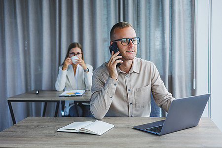 男性经理在用电脑和笔记板坐在一张桌子上时 正在用移动电话说话 在有大窗户的办公室里工作气氛良好人士领导照片互联网技术团队成人男人图片