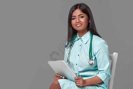 身着水生洋装的听诊器 坐在灰底椅子上的印度女医生护士 她配戴耳眼透镜医疗保健帮助药品专家卫生蓝晶保险诊断职业工作背景图片