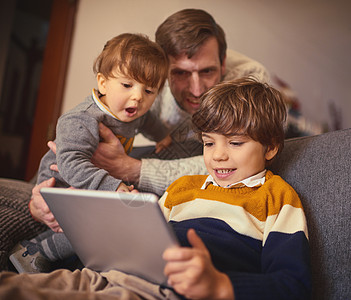 让我们看看你的兄弟们在做什么 一个可爱的小男孩在家里的沙发上使用平板电脑 而他的父亲和弟弟则在一旁看着图片