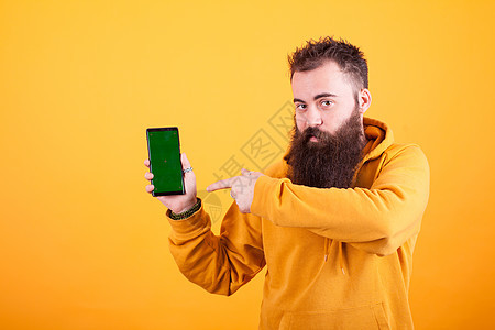 在黄色背景上指着智能手机的 迷人的胡须嬉马男子成人人士学生工人商务手势帽衫快乐手指潮人图片