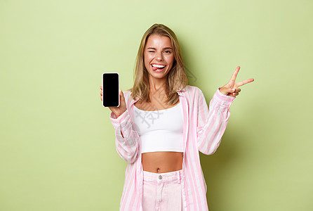 穿着粉红色衬衫的金发美女肖像 展示和平标志和手机屏幕 微笑喜悦 站在绿色背景上站立图片