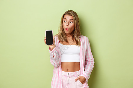 粉红色衬衫中金发美女女性模特的图像 显示智能手机屏幕和惊异的外表 站在绿色背景之上图片