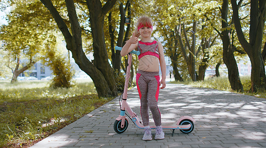 运动健壮的年轻女孩骑着电动摩托车 站在自行车旁边看公园中的摄像机教育慢跑者享受训练孩子们赛跑者青少年速度乐趣女学生图片