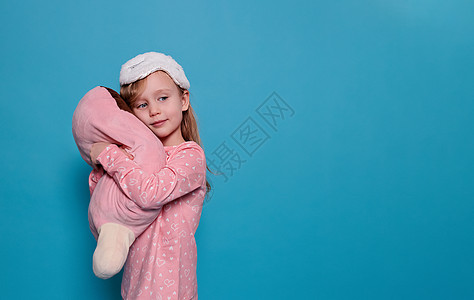 穿着睡衣和睡眠面具的小女孩拥抱着蓝色背景的玩具 复制空间图片
