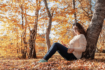 年轻有见识的女人坐在秋林的树旁女士季节健康森林毛衣公园闲暇橙子头发成人图片