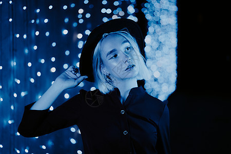 千禧年神秘漂亮的女孩金发发型在晚上靠近蓝色发光的霓虹灯墙 头发 时髦的帽子 鼻子穿孔 神秘女子短信应用成人游客商务衣领手机繁荣享图片