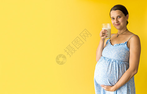 肖像可爱美丽怀孕的年轻漂亮的女人在花卉蓝色连衣裙拿着喝杯牛奶在黄色背景上揉肚子 母性 女性气质 健康饮食 乳制品 夏季概念保健女图片