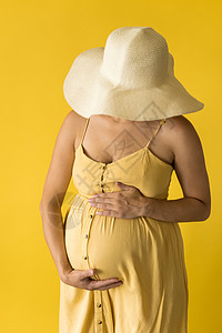 母性 女性气质 时尚 炎热的夏天 — 穿着漂亮复古连衣裙的特写镜头无法辨认的孕妇女士普罗旺斯草帽 双手放在肚子上 在黄色背景上揉图片