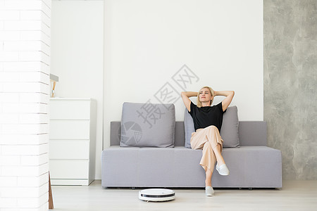 使用自动真空吸尘器清洁地板 控制机器家务机器人的年轻女性公寓房子微笑广告卫生黑发技术清扫沙发家政图片