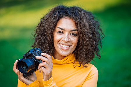 身穿黄衣的年轻非洲美女用DSLR相机拍摄绿色背景照片 女孩笑得像摄影师一样开心公共公园爱好女人游客休闲成人乐趣微笑女性活动图片