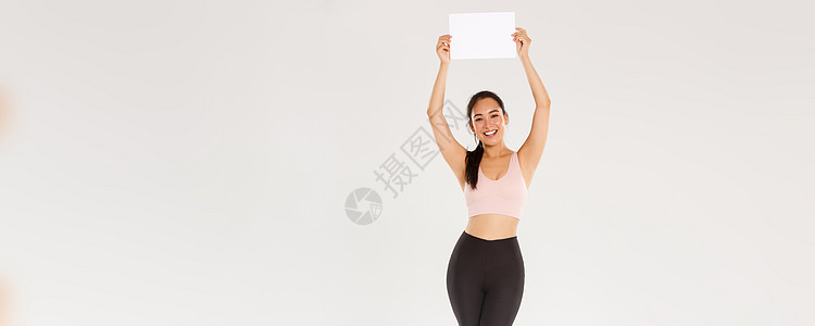 全长欢快 苗条 快乐的可爱女孩在健身房训练 穿着运动服 举着牌子 一张空纸 上面有你的广告 站在白色背景讲师身体运动装跑步健美教图片