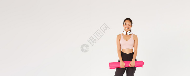 全长微笑苗条的亚洲女孩 带着橡胶垫进行健身锻炼 戴着耳机 在等待训练教练开始在健身房锻炼时看着相机 白色背景广告女性讲师身体运动图片