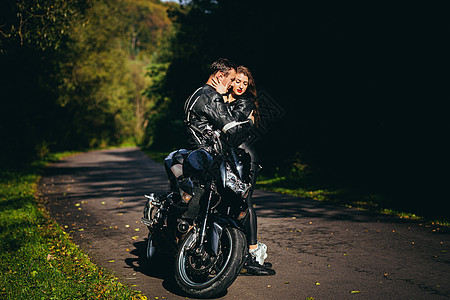 英俊的年轻男子和美丽的年轻女孩 一对夫妇坐在摩托车上 站在一辆穿着黑色皮衣的摩托车旁边 在大自然中拥抱 在街上假期幸福运输公路活图片
