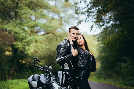 英俊的年轻男子和美丽的年轻女孩 一对夫妇坐在摩托车上 站在一辆穿着黑色皮衣的摩托车旁边 在大自然中拥抱 在街上男人假期运输女朋友图片