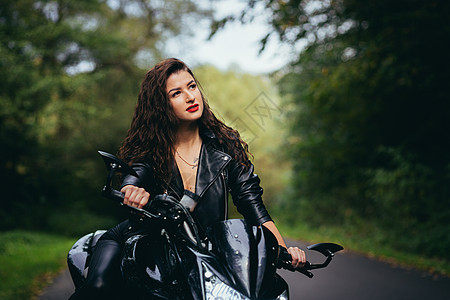 一位穿着黑色皮衣骑摩托车的年轻美女的肖像 在自然 街道 森林 阳光明媚的天气里 闭着眼看摄像机车辆棕色模特气氛时装目的地方式运输图片