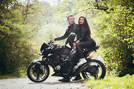 英俊的年轻男子和美丽的年轻女孩 一对夫妇坐在摩托车上 站在一辆穿着黑色皮衣的摩托车旁边 在大自然中拥抱 在街上活动乐趣公路服装皮图片