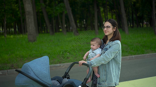 一位年轻的母亲抱着一个六个月大的儿子走路 公园里 一位戴眼镜的女士背着婴儿 推着婴儿车图片