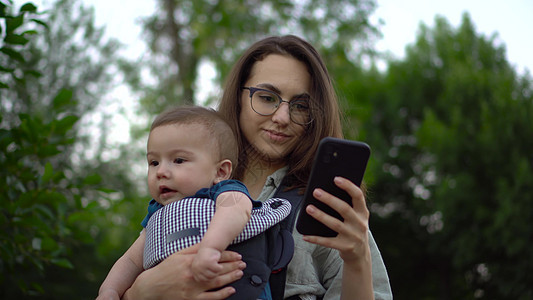 一位带着孩子的年轻女子站在公园里 手里拿着电话 戴眼镜的妈妈抱着儿子 袋鼠背包里的新生儿子 特写图片