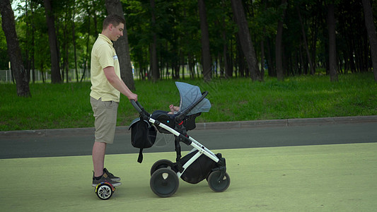 一个年轻人骑着陀螺滑板车 推着婴儿车 现代父亲带着孩子在公园散步 黄色 T 恤和浅色短裤图片