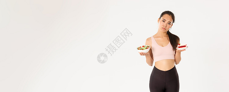 积极的生活方式 健身和幸福理念 忧郁而悲伤的可爱亚洲女运动员拿着美味的蛋糕和绿色沙拉 生闷气 因为不能吃甜食 节食 站在白色背景图片