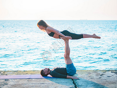 在海边做Acroyoga的年轻夫妇 男人躺在混凝土板上 让女人靠在脚上平衡 漂亮的一对一起练瑜伽练习朋友们花园女性冒充力量运动身图片