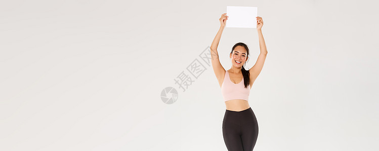 全长欢快 苗条 快乐的可爱女孩在健身房训练 穿着运动服 举着牌子 一张空纸 上面有你的广告 站在白色背景健美讲师肌肉促销跑步福利图片