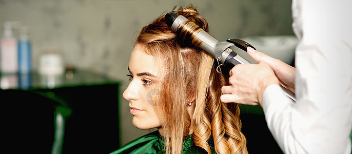 发型师用卷纹长棕色头发 在美容院里 对着年轻的天主教女孩魅力理发理发师女性黑发造型师发型设计造型卷发器工作图片