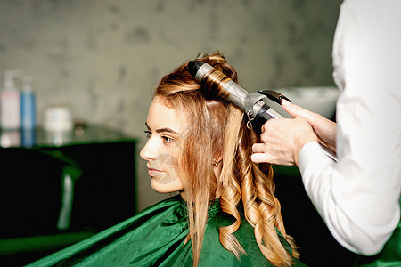 发型师用卷纹长棕色头发 在美容院里 对着年轻的天主教女孩新娘工作室电气卷发器发型设计造型商业发型身体婚礼图片