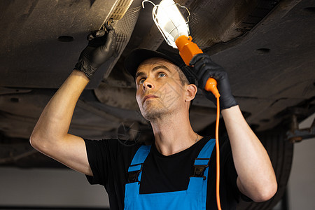 白人汽车修理工站着 LED 灯 看着汽车零件 在大修时擦拭汽车下方的发动机 以换油并维护车库中的发动机图片