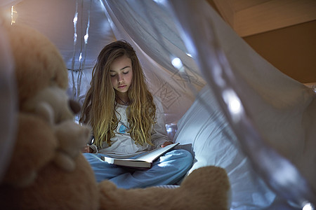 一个年轻女孩在家里的毯子堡下 读着一本书 她很会写东西图片
