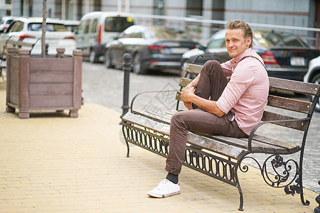英俊的年轻男子坐在长椅上 一脚踩在凳子上 在城市办公室或家附近度过时间 穿着粉红色衬衫和肩膀袋图片