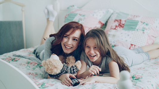 欢笑快乐的母亲和可爱的女儿在电视上看滑稽卡通电影时 早上躺在家里的床上 玩得开心公寓妈妈闲暇孩子寝具睡衣卧室家庭微笑金发女郎图片