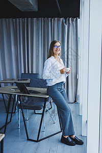 年轻微笑的女商务人士站在靠近窗户的办公室里 手里拿着咖啡 一位戴眼镜和白衬衫的女经理正在下班休息杯子工人笔记本眼镜自由职业者技术图片