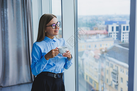 一个穿着经典衬衫和眼镜的快乐年轻女人的形象 她站在有大窗户的现代办公室的窗边微笑着喝着咖啡 远程工作医生互联网杯子经理技术女士饮图片