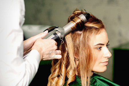 发型师用卷纹长棕色头发 在美容院里 对着年轻的天主教女孩工作室造型治疗电气美容师女性卷发器沙龙梳妆台造型师图片