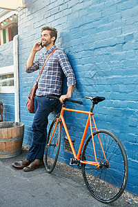一位英俊的年轻人靠在墙上 骑着自行车用手机说话 他把车停在墙边图片