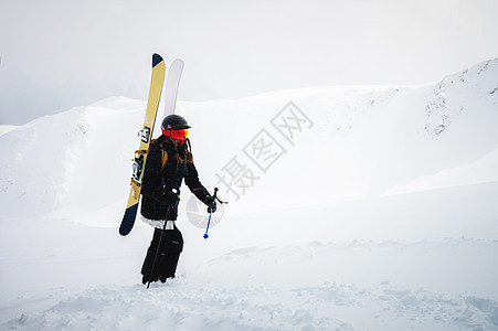 带着滑雪设备和棍子的年轻女子正在攀登偏远地区的雪山 有雾的天空和多雪的山在背景中图片