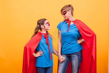 母亲和女儿穿得像超级英雄 手牵手 仰望着对方 在黄色背景上快乐航班父母动机拥抱戏服喜悦童年力量乐趣图片