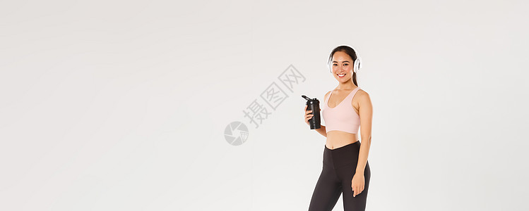 全长微笑苗条和快乐的健身女孩 亚洲女性运动员在健身房锻炼 戴着耳机 在锻炼期间听音乐 在锻炼之间从瓶子里喝水 白色背景身体促销教图片
