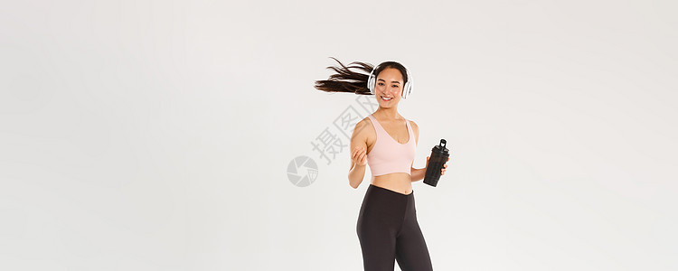 全长微笑迷人的亚洲女性运动员 在健身房锻炼身体的女孩 女运动员或跑步者从瓶子里喝水 用无线耳机听音乐 甩头发健美情绪促销教练训练图片