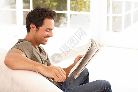 这篇文章很有趣 一个英俊的年轻人坐在沙发上 在家看报纸的时候坐着图片