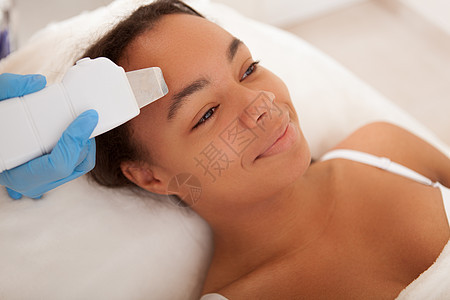 接受面部治疗的非洲青年妇女沙龙护理医生仪器毛孔机器美容皮肤科职业顾客图片